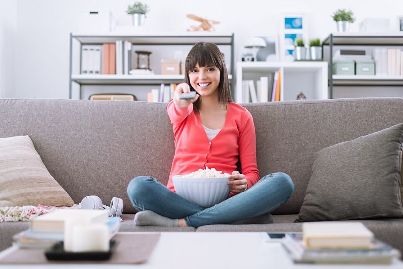 Woman sitting on sofa with popcorn and remote control, enjoying streaming services in Australia